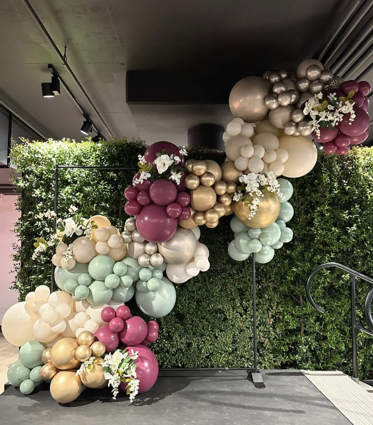 Blush, gold, and mint balloon arch against a greenery wall